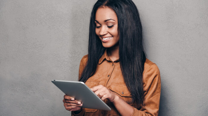 Beauty with digital tablet. Beautiful young African woman holding touchpad and looking at it with smile while standing against grey background