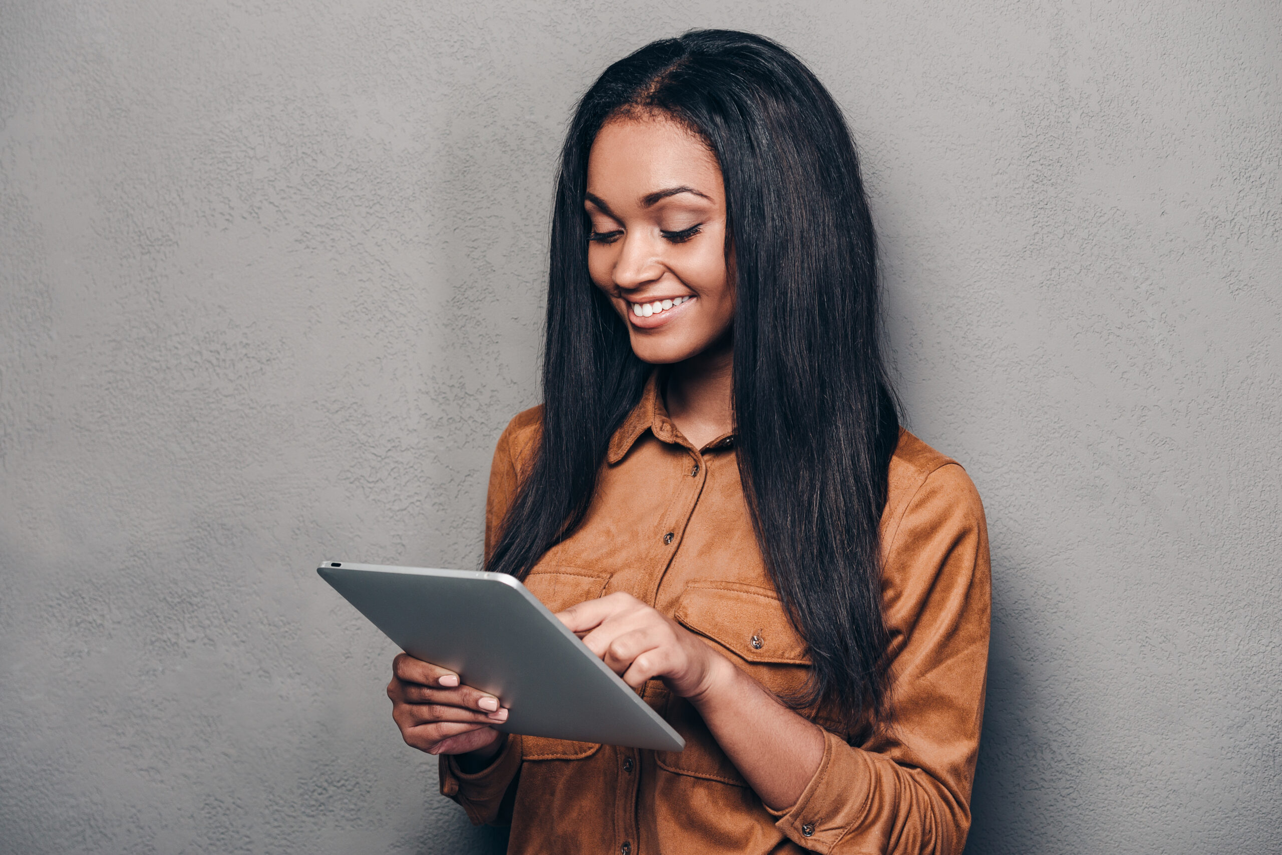 Beauty With Digital Tablet. Beautiful Young African Woman Holding Touchpad And Looking At It With Smile While Standing Against Grey Background