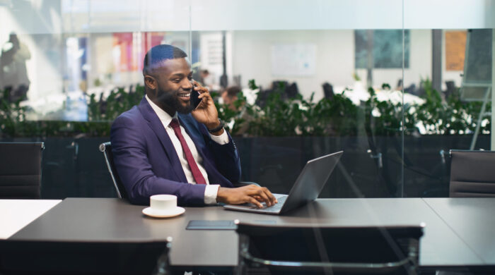 Cheerful young black businessman working on laptop at office, having phone conversation with his personal assistant or business partner and smiling, panorama with copy space, shot through glass walls