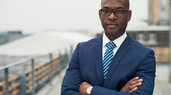 Confident black business man in a stylish suit standing with folded arms on a rooftop of n office block looking at the camera with a serious expression