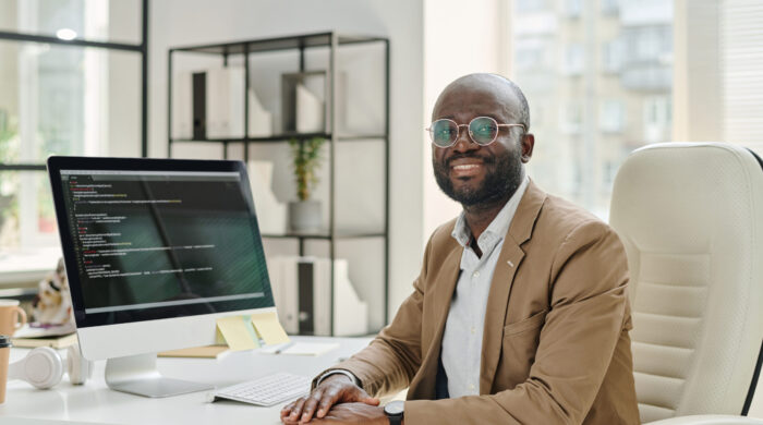 Portrait of African developer in eyeglasses smiling at camera while sitting at his workplace with computer monitor with codes on screen