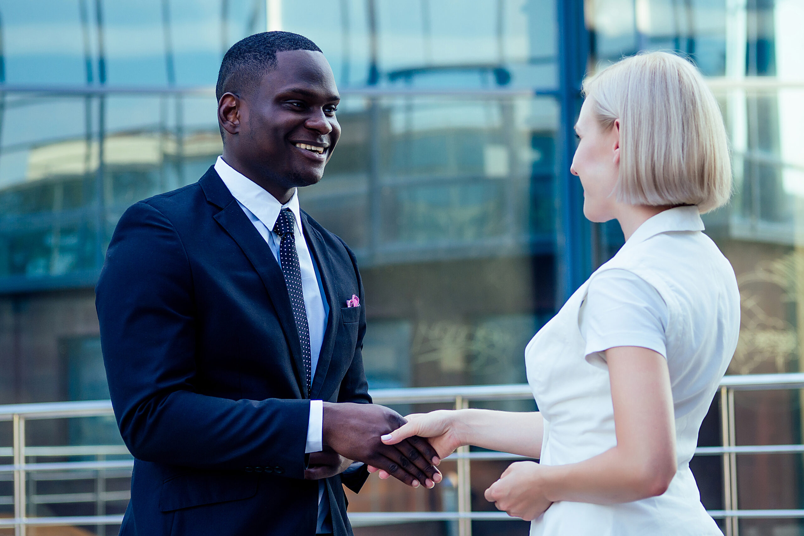 Handsome African American Man In A Black Business Suit Shaking Hand With A Businesswoman Partner Cityscape Glass Offices Background. Teamwork And Successful Deal Idea