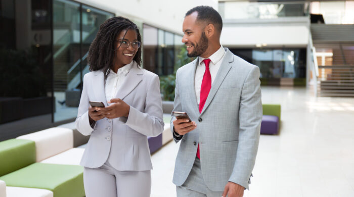 Happy business colleagues using mobile phones and chatting. Business man and woman walking through office hallway, holding smartphones and talking. Corporate communication concept