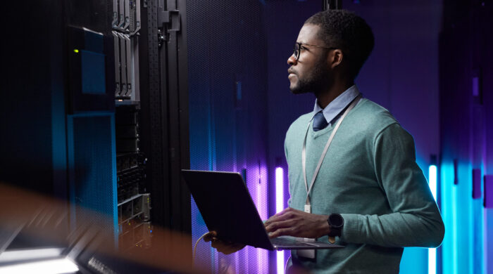 Side view portrait of African American data engineer holding laptop while working with supercomputer in server room lit by blue light, copy space