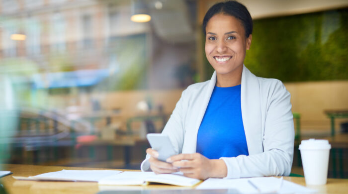 Cheerful young manager in smart casual sitting by table, messaging in smartphone and relaxing at coffee-break