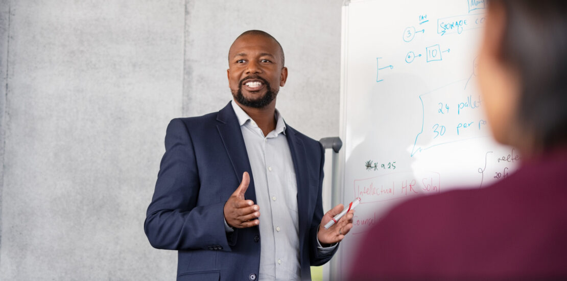 Mature african american coach explaining strategy to his team with copy space. Successful businessman presenting new project to employees during meeting room. Conference speaker talk to audience while giving presentation on whiteboard to business group of people.