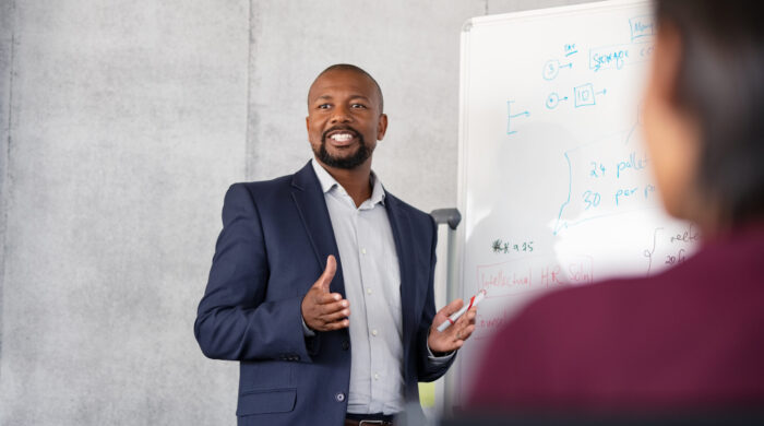 Mature african american coach explaining strategy to his team with copy space. Successful businessman presenting new project to employees during meeting room. Conference speaker talk to audience while giving presentation on whiteboard to business group of people.