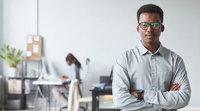 Waist up portrait of successful African-American man looking at camera while standing with arms crossed in office, copy space