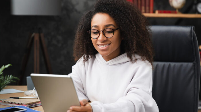 Smiling cheerful african woman teenage girl schoolgirl using digital tablet at home, doing homework assignment online, browsing social media