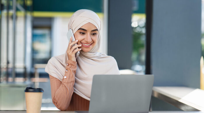 Workplace outdoor, modern independent contractor or business woman work in city with tech. Smiling young arab lady in hijab speaks on phone and looks at laptop, at table with cup of coffee, copy space