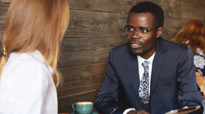 Young African businessman holding tablet and sharing data with female Caucasian colleague during coffee break in cosy caf�Smart coworker makes an eye-contact with job partner and listens to her.