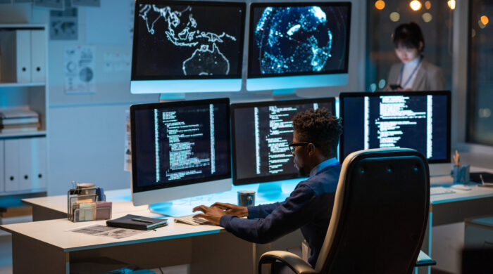 Young contemporary cyber security manager typing while sitting by desk in front of computer monitors