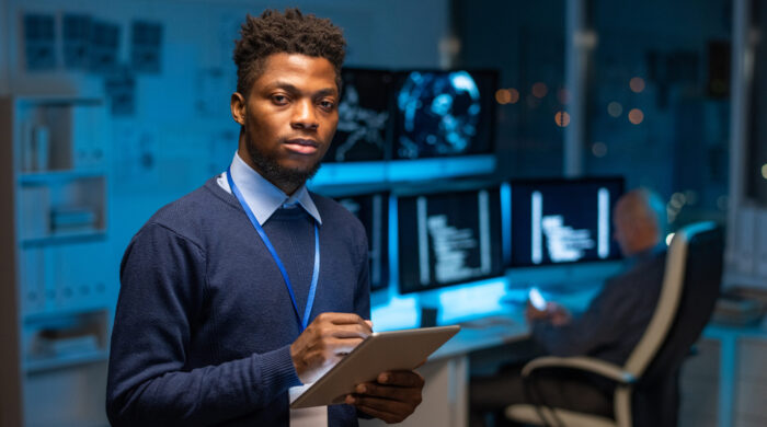 Young serious African programmer with tablet standing in front of camera against colleague by workplace